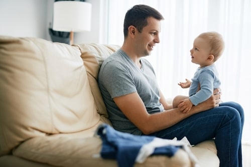 father sits on couch with baby on lap, infant is gesturing and gazing into father's eyes