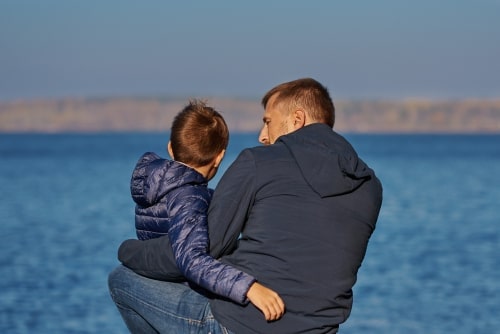 father sits on the shore with young boy on his lap, talking to son 