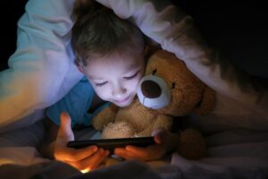 young child under covers with teddy bear, holding electronic media device
