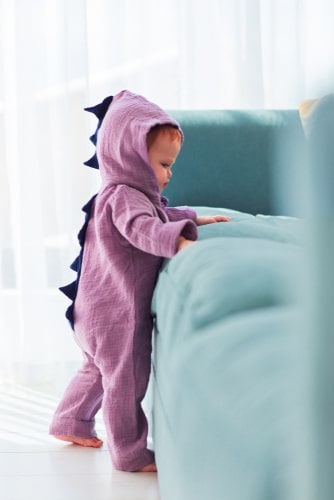 cruising baby, wearing dinosaur pajamas, is standing up and holding onto a sofa to move around