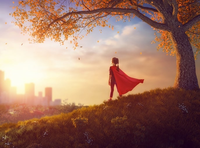 little girl, wearing a superhero cape, is standing on a hill, looking down at a city.