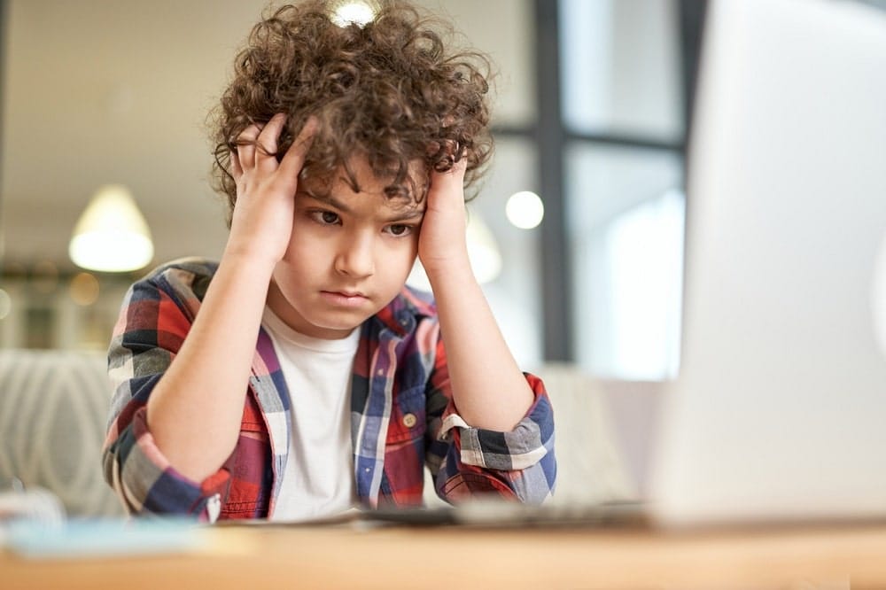 tired, sleep deprived young boy holding head in hands while looking at computer monitor.