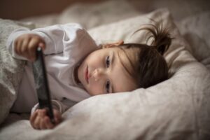 little girl in bed, holding and examining a hand-held electronic device