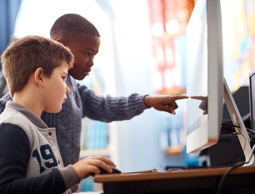 two boys working on a computer together, one boy using a mouse while the other boy points to the screen