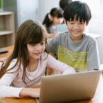 happy, primary school children in classroom - girl on laptop with boy watching