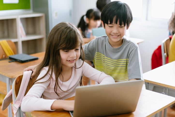 happy, primary school children in classroom - girl on laptop with boy watching