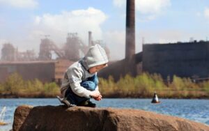 toddler crouched on a rock outdoors, with a factory in the distance - air pollution