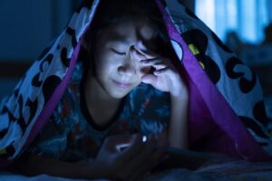 young girl awake in bed at night, rubbing her eyes while looking at mobile phone screen