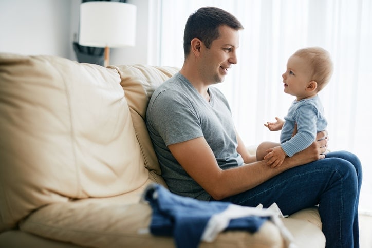 baby sits on father's lap; they are talking happily, face to face, and baby is gesturing