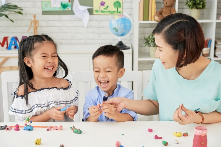 mother sits at table with two laughing children as they make things with clay