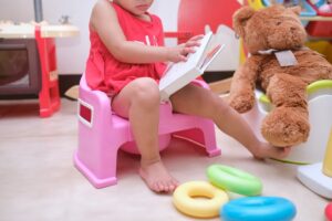 Young child on potty, viewed from the shoulders down, reading a book and sitting next to a teddy bear on another potty