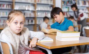 girl in library at desk gazes at viewer, while boy studies in the background