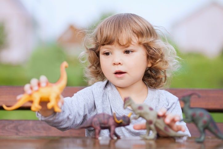toddler girl playing outside with four different plastic dinosaur toys, including a sauropod, Pachycephalosaurus, and hadrosaur