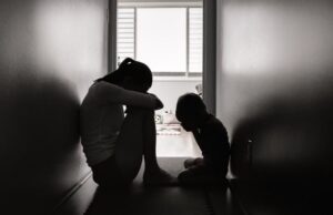 grey silhouette of mother and child sitting in dark hallway, each looking down in despair