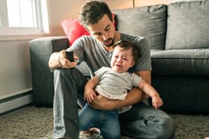 father holds distressed infant while both look at a TV screen