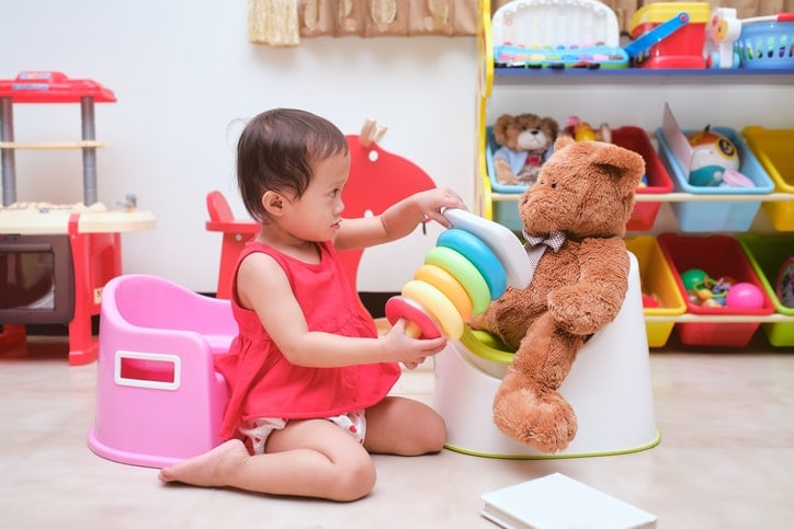 toddler girl offers a toy to a teddy bear on a potty chair