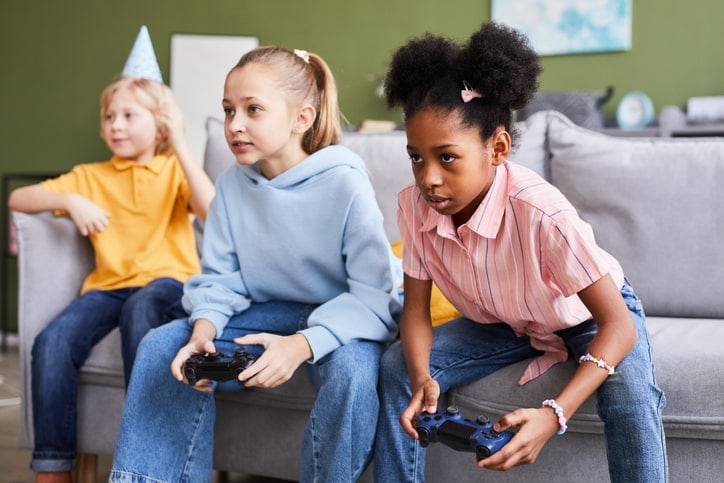 two girls playing video games intensely on a couch, while boy watches