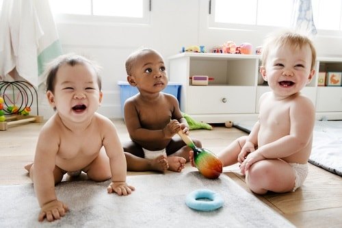 Three babies, different races, sitting together on the floor of a playroom.