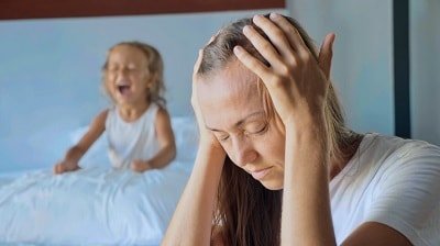 stressed out mother holds her head in her hands while her young daughter throws tantrum in the background