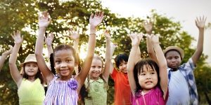 diverse assortment of young children, representing different races, cheerfully raising their hands outside