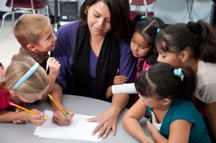 Kids benefiting from quality student-teacher relationships: Young children surrounding their teacher while she demonstrates something on paper. 