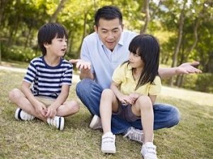 father sits on grass with daughter and son, engaged in friendly talk, gesturing with hands